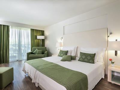 Bright hotel room with double bed, green accents, sofa, and balcony with sheer curtains.