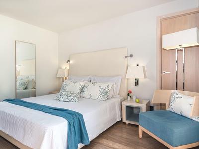 Bright hotel room with large bed, teal accents, wooden floor, and a small armchair.