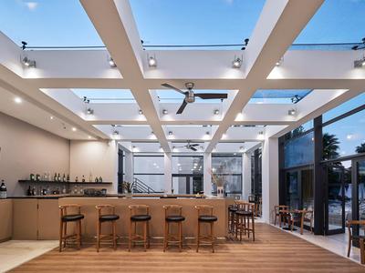 Moderní hotelový bar s dřevěným pultem, barovými židlemi a velkými skleněnými stěnami pod otevřenou oblohou.