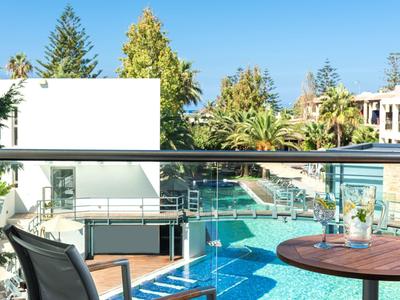 Modern outdoor pool area with sun loungers, glass railing, and greenery under blue sky.
