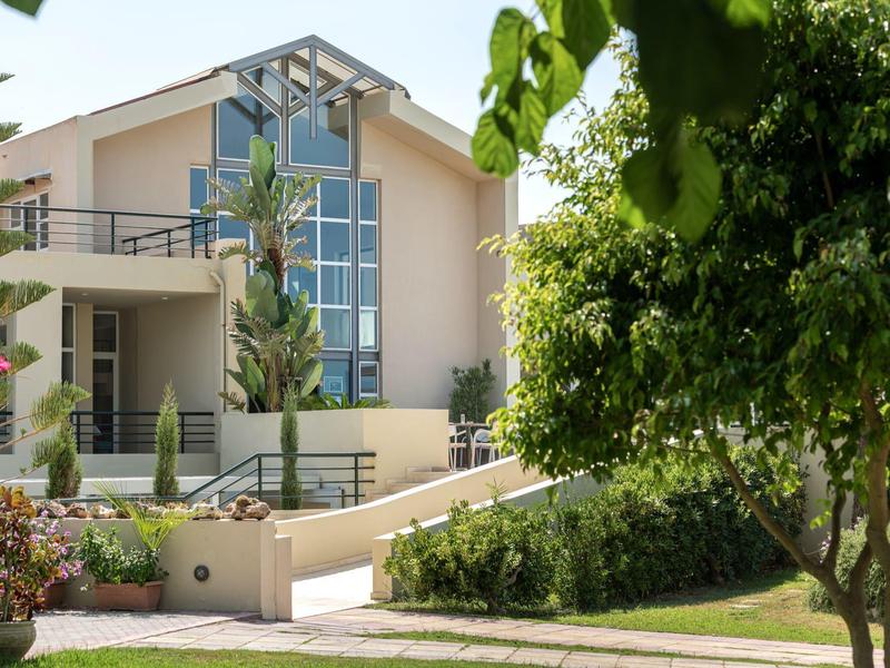 Modern beige villa with large windows surrounded by lush greenery and palm trees.