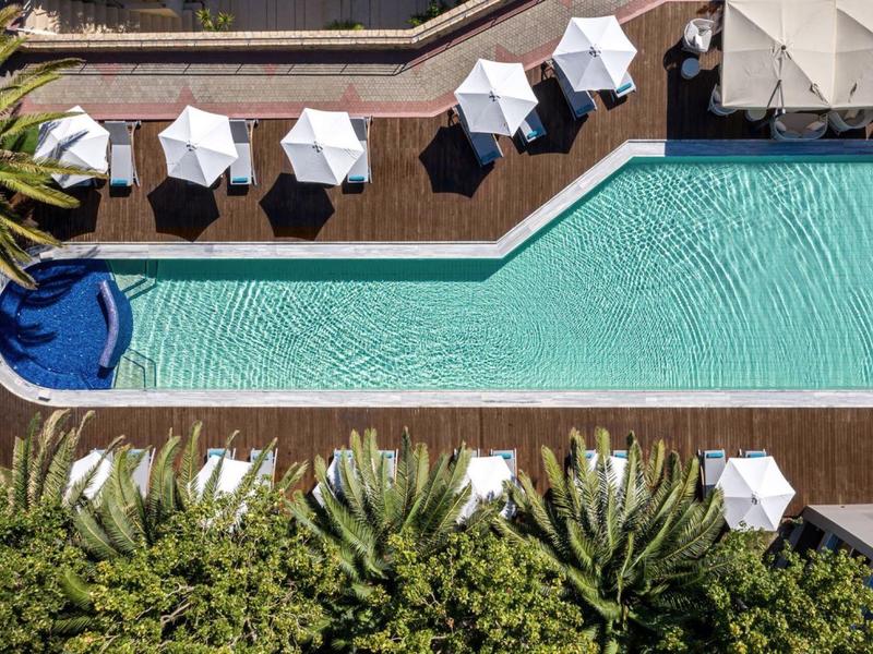 Overhead view of a pool with white umbrellas, sun loungers, and greenery around a wooden deck.