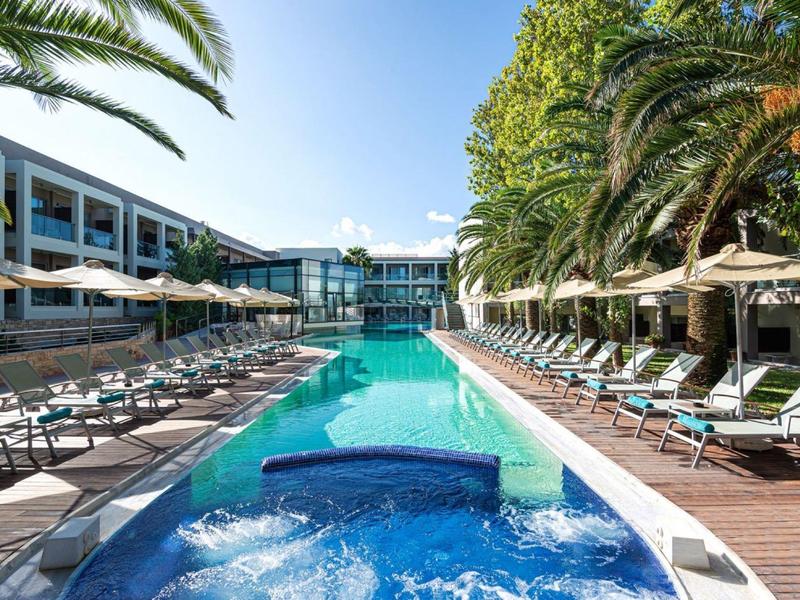 Outdoor pool area at a hotel with loungers, umbrellas, and palm trees under a clear sky.