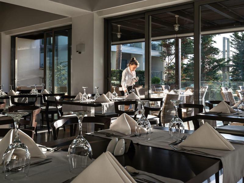 Elegant hotel restaurant with table settings and a server preparing a table near large windows.