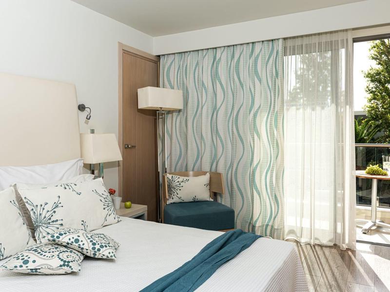 Bright hotel room with queen bed, patterned curtains, armchair, and balcony with outdoor seating.