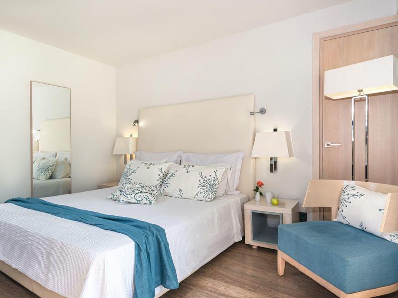 Bright hotel room with large bed, teal accents, wooden floor, and a small armchair.