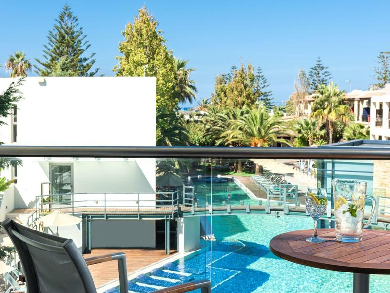 Modern outdoor pool area with sun loungers, glass railing, and greenery under blue sky.