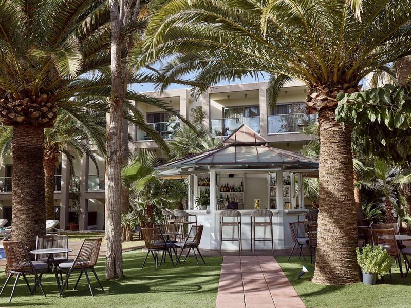 Outdoor hotel bar with palm trees, seating area, and green lawn under blue sky.