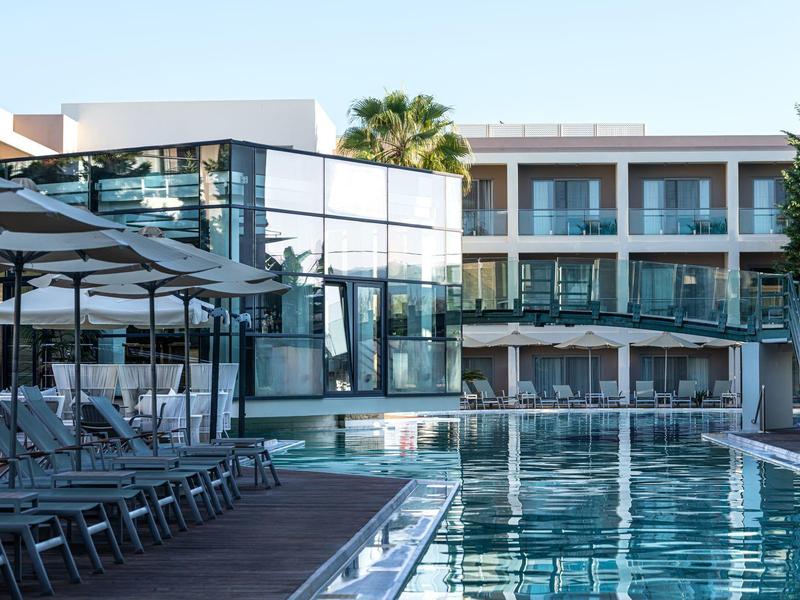 Modern hotel with glass facade, pool, sun loungers, and umbrellas under clear sky.