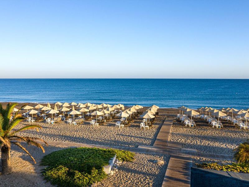 Sandy beach with rows of sun loungers and umbrellas by the calm blue sea under clear sky.
