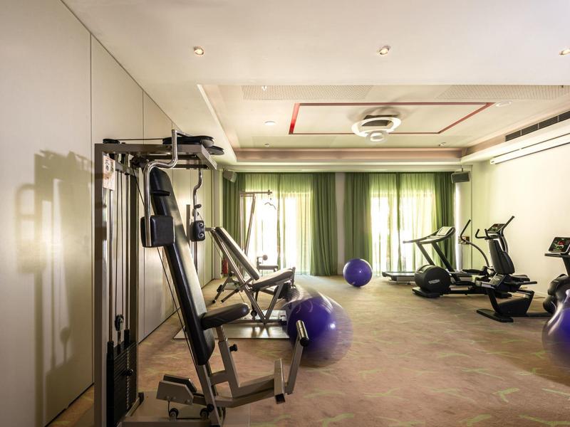 Hotel gym with various exercise machines and large windows with green curtains.