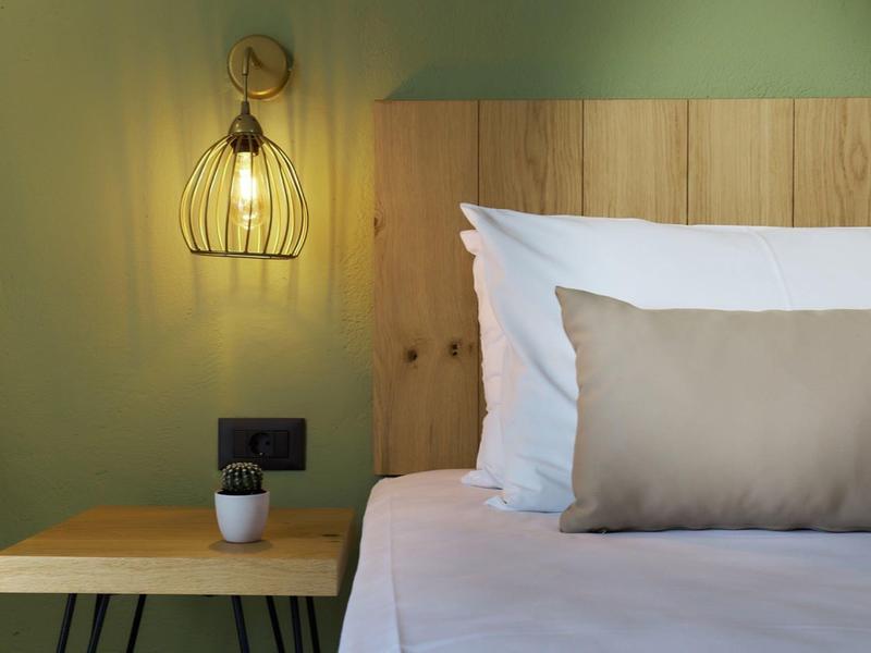 Przytulny pokój hotelowy z drewnianym zagłówkiem łóżka, stolikiem nocnym i nowoczesną lampą ścienną.