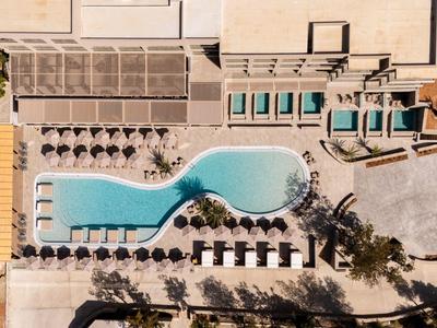 Vista aerea di una piscina curva con lettini e ombrelloni accanto a un hotel.