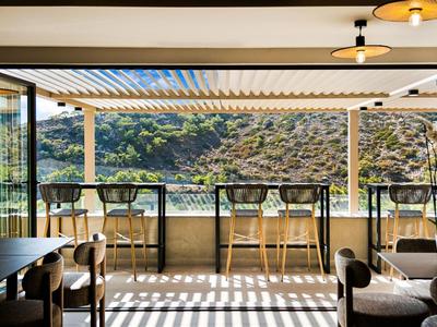 Een overdekt buitenterras met hoge tafels en stoelen met uitzicht op heuvelachtig landschap en water.