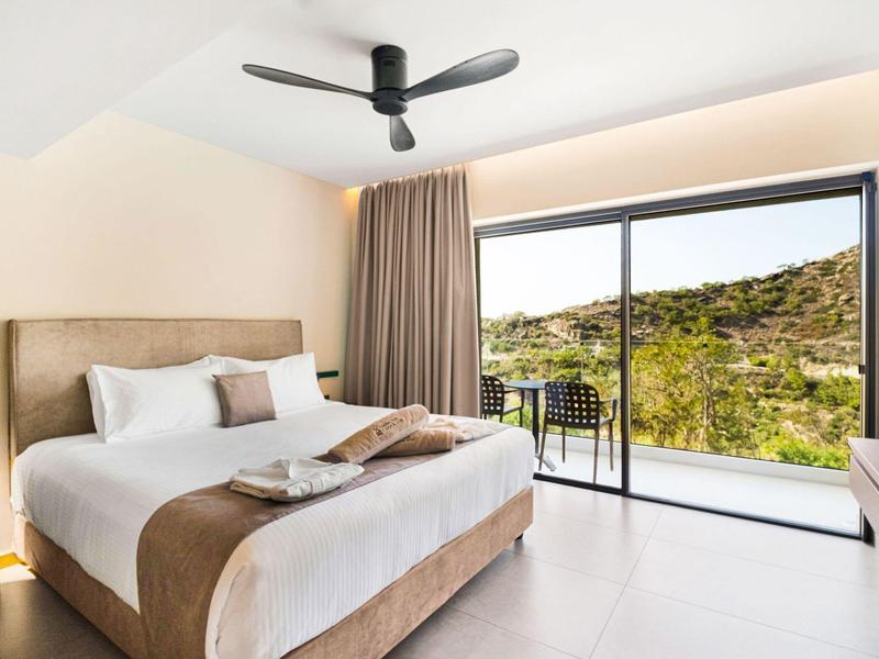 Moderne hotelkamer met groot bed, plafondventilator en balkon met uitzicht op heuvels.