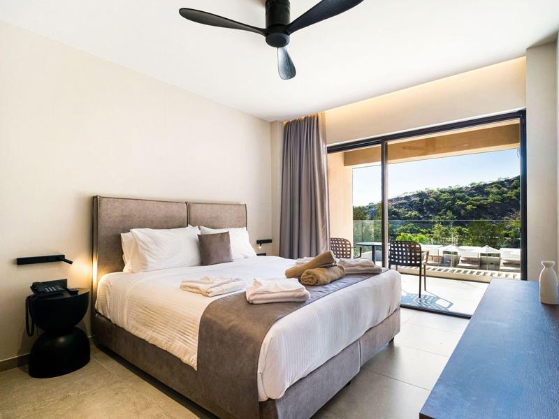 Lichte hotelkamer met tweepersoonsbed, plafondventilator en balkon met uitzicht op groene heuvels.