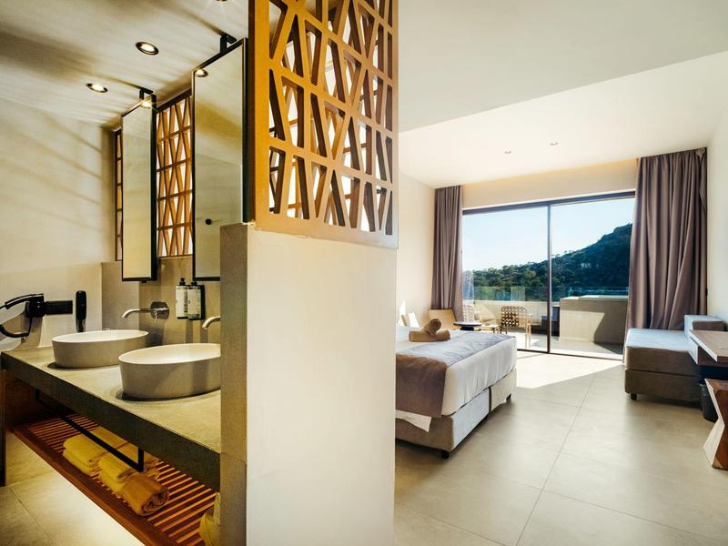 Licht hotelkamer met groot raam, bergzicht en moderne badkamer met twee wastafels.