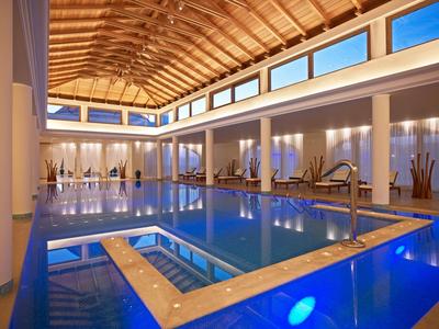 Modernes Hallenbad mit großem Schwimmbecken, Holzbalkendecke und hellen Wänden, Tageslicht fällt ein.