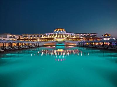 Beleuchtetes Luxushotel mit großem Pool bei Nacht, klare Reflexion im ruhigen Wasser.