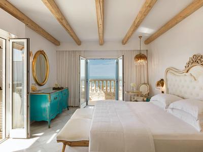 Helles Schlafzimmer mit weißen Wänden, Holzbalken, Doppelbett und Meerblick durch offene Türen.