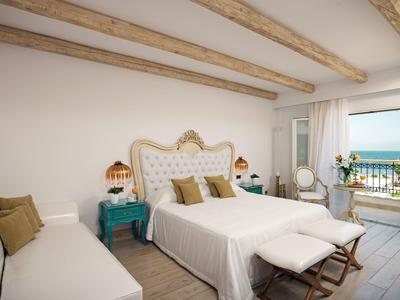 Helles Schlafzimmer mit weißem Bett, Holzdeckenbalken, zwei Nachttischen und Meerblick vom Balkon.