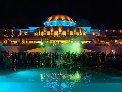 Beleuchtetes Hotel mit kuppelförmigem Dach und Pool, vor dem sich Menschen versammeln.