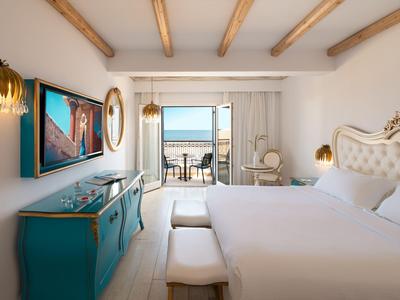 Moderní hotelový pokoj s balkonem a výhledem na moře, světlý s bílým vybavením a dřevěnými stropy.