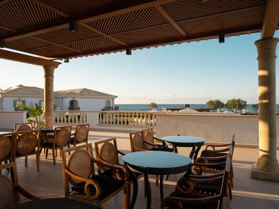 Zastřešená terasa se stoly a židlemi s výhledem na budovy a moře.