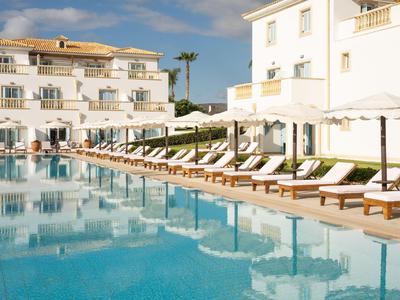 Oblast bazénu s mnoha lehátky a bílými slunečníky před vícepodlažními hotelovými budovami.