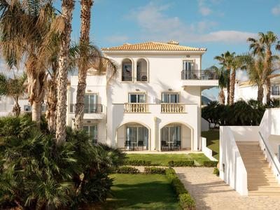 Bílý rekreační dům s balkonem, obklopený palmami a zahradou pod modrou oblohou.