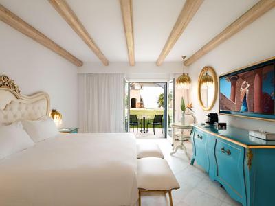 Světle hotelový pokoj s velkou postelí, tyrkysovou komodou a výhledem na zahradu a bazén.