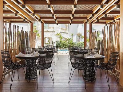 Otevřená restaurace s dřevěným stropem, černými drátěnými židlemi a výhledem na bazén.