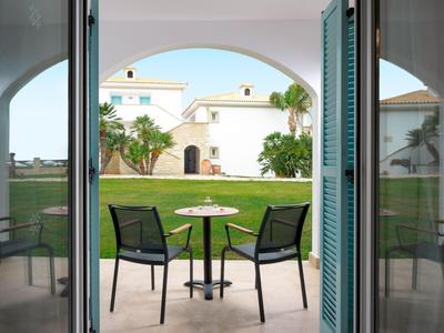 Pohled skrz otevřené dveře na terasu s stolem, židlemi a zahradou před bílou budovou.