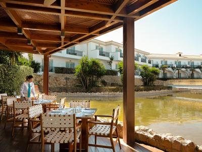 Zastřešená terasa s prostřenými stoly vedle rybníka před hotelovým komplexem za slunečného dne.