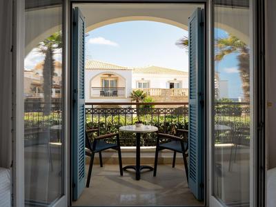 Balkon se dvěma židlemi a stolem s výhledem na hotelovou budovu a palmy.