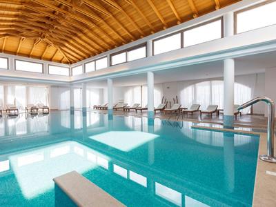 Vnitřní bazén s velkou plochou vody, lehátky a vysokým stropem s dřevěnými trámy.