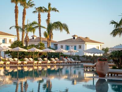Hotelpool mit Sonnenliegen, Sonnenschirmen und Palmen vor weißen Gebäuden bei klarem Himmel.