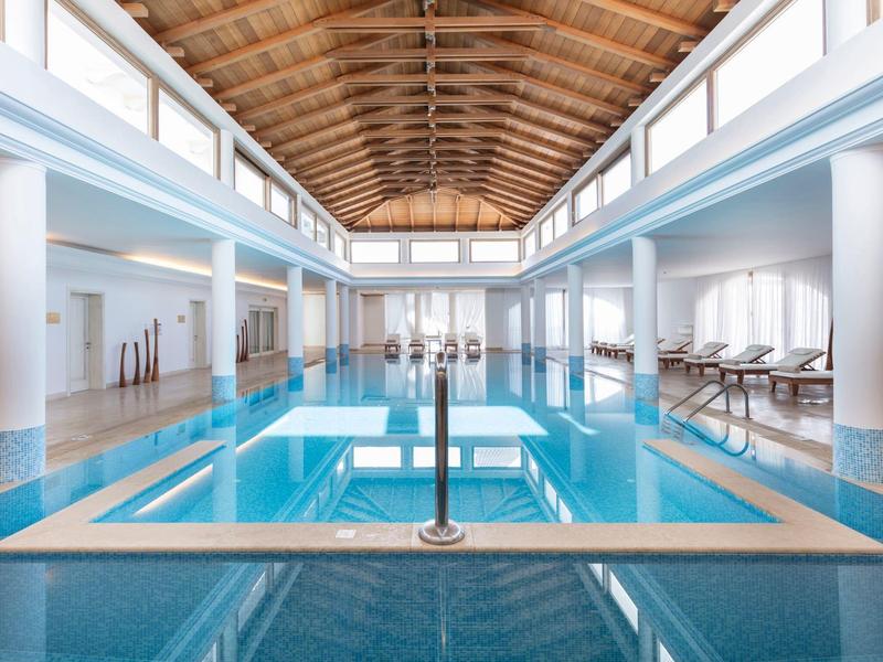Piscina interna con soffitto in legno e lettini in un'area benessere luminosa