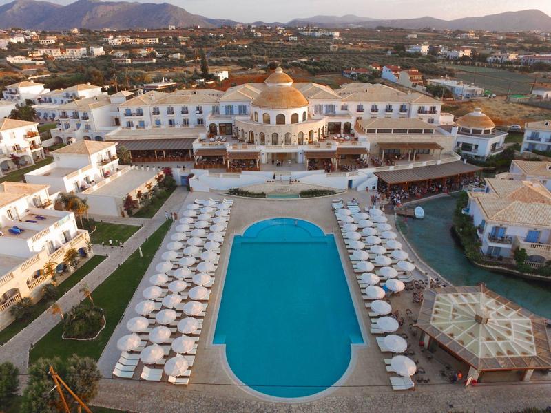 Grande hotel con piscina ovale e ombrelloni davanti a un paesaggio montano.