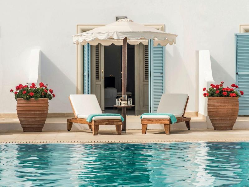 Due lettini con un ombrellone tra di loro accanto alla piscina davanti a un muro bianco con porte blu.