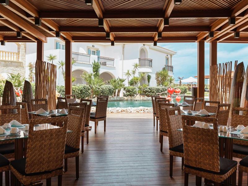 Ristorante aperto con tetto in legno che si affaccia su una piscina e palme in una giornata di sole.