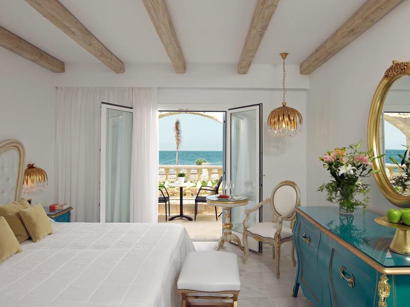 Camera luminosa con biancheria bianca, travi in legno, credenza blu e vista mare dal balcone.