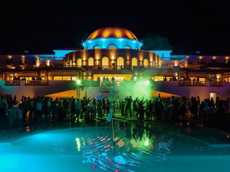 Hotel illuminato di notte con piscina e persone in primo piano.