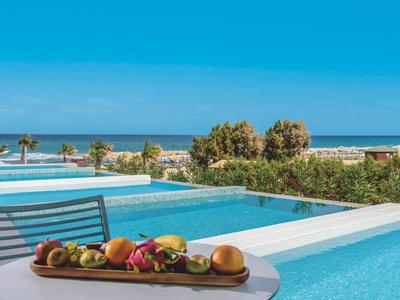 Pool mit klarem Wasser, Tisch mit Obst und Blick auf Meer und Palmen im Sonnenschein.