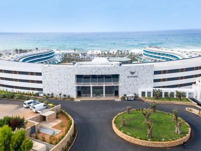 Großes rundes modernes Gebäude mit weißer Fassade, blauen Glasfenstern und Meer im Hintergrund.