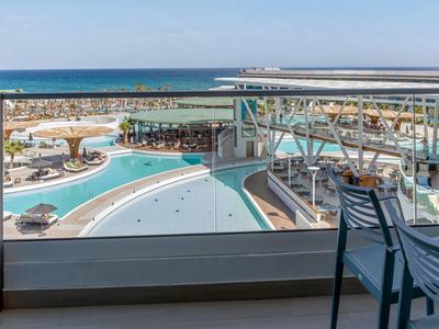 Balkon mit Stuhl blickt auf großen Pool, Strand und blaues Meer unter klarem Himmel.