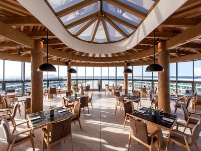 Moderner, heller Restaurantraum mit Holzsäulen, großen Fenstern und Panoramaausblick auf Wasser und Himmel.