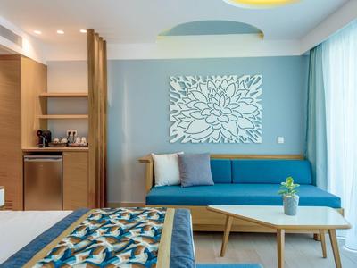 Modernes Hotelzimmer mit blauem Sofa, Holzregal, Bett mit gemustertem Überwurf und großem Fenster.