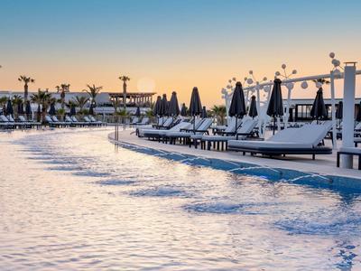 Großer Pool bei Sonnenuntergang mit Liegestühlen und Sonnenschirmen am Rand, Palmen im Hintergrund.