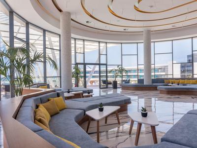 Modernes, helles Lounge mit großen Fenstern, grauen Sofas, runden Tischen und Pflanzen.